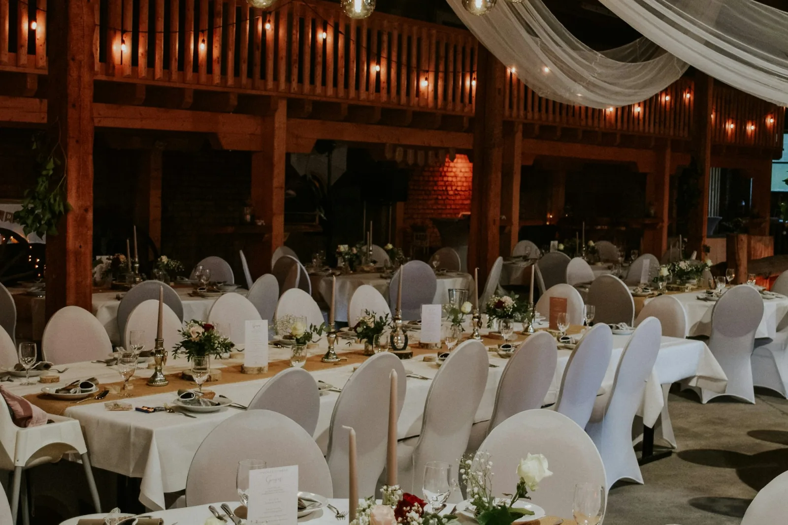 Sảnh tiệc bày bàn dài phủ khăn trắng, ghế bọc trắng, rèm voan và dây đèn trên trần