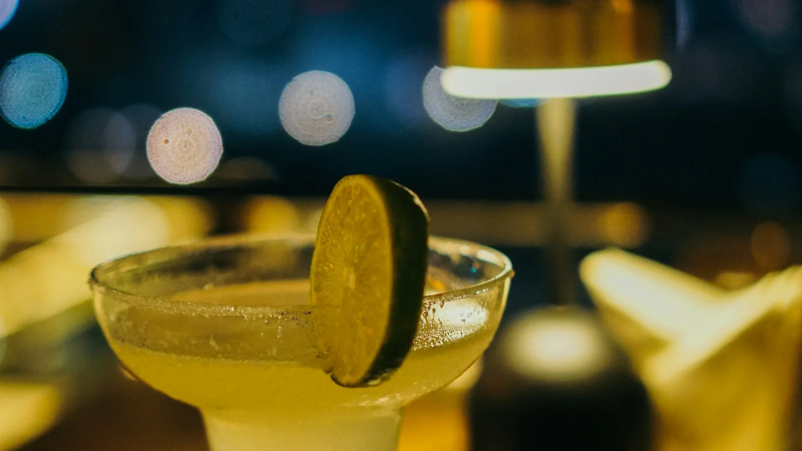 Ly cocktail chanh và đèn bàn trên quầy bar buổi tối, phía sau là ánh đèn thành phố nhòe